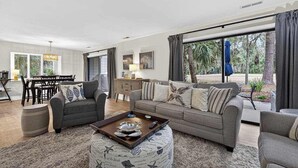 A spacious living room with a sofa, ottoman, and coffee table, a dining area with a table and chairs, and large windows with curtains. - Charming Townhome with Golf Course View,On-site Pool and Short Walk to Beach! (Hilton Head)