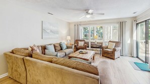 Ein Wohnzimmer mit einem Ecksofa, Sesseln, einem Couchtisch, einem Deckenventilator und großen Fenstern.