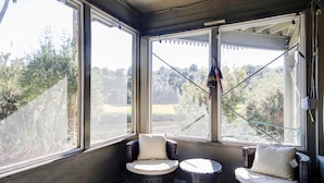 Ein Wintergarten mit zwei Korbstühlen und einem kleinen Tisch, große Fenster mit Blick auf Bäume und einen Zaun.