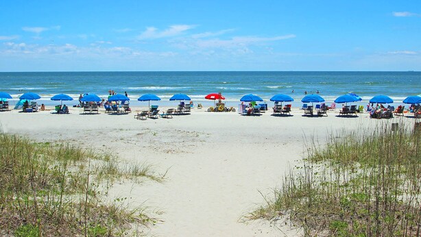 Ein Strand mit Reihen blauer Sonnenschirme und Liegestühle, ein klarer Himmel und das Meer im Hintergrund.