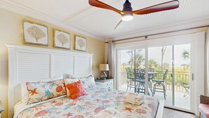 Ein Schlafzimmer mit Bett, Deckenventilator, Nachttisch, Lampe, Fenster und Balkon mit Sitzgelegenheiten.