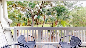 A covered porch with two wicker chairs and a small table, overlooking a lush garden with various trees and shrubs. - Driftwood Villa - 261 | Golf course-view condo with balcony, W/D, AC - dogs OK (Edisto Beach)