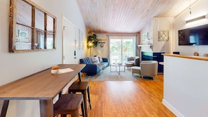 A spacious living area with a wooden ceiling, a dining table, a sofa, and a TV mounted on the wall. - Driftwood Villa - 261 | Golf course-view condo with balcony, W/D, AC - dogs OK (Edisto Beach)
