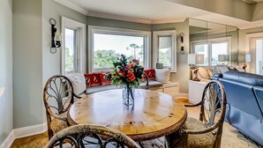 A dining area with a round wooden table, chairs, a vase of flowers, and a view through large windows. - Shorewood, Unit 530 Penthouse condo in Hilton Head with ocean views (Hilton Head)