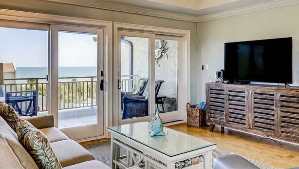 A living room with a sofa, a glass coffee table, a television on a wooden cabinet, and a balcony with a view of the sea. - Shorewood, Unit 530 Penthouse condo in Hilton Head with ocean views (Hilton Head)