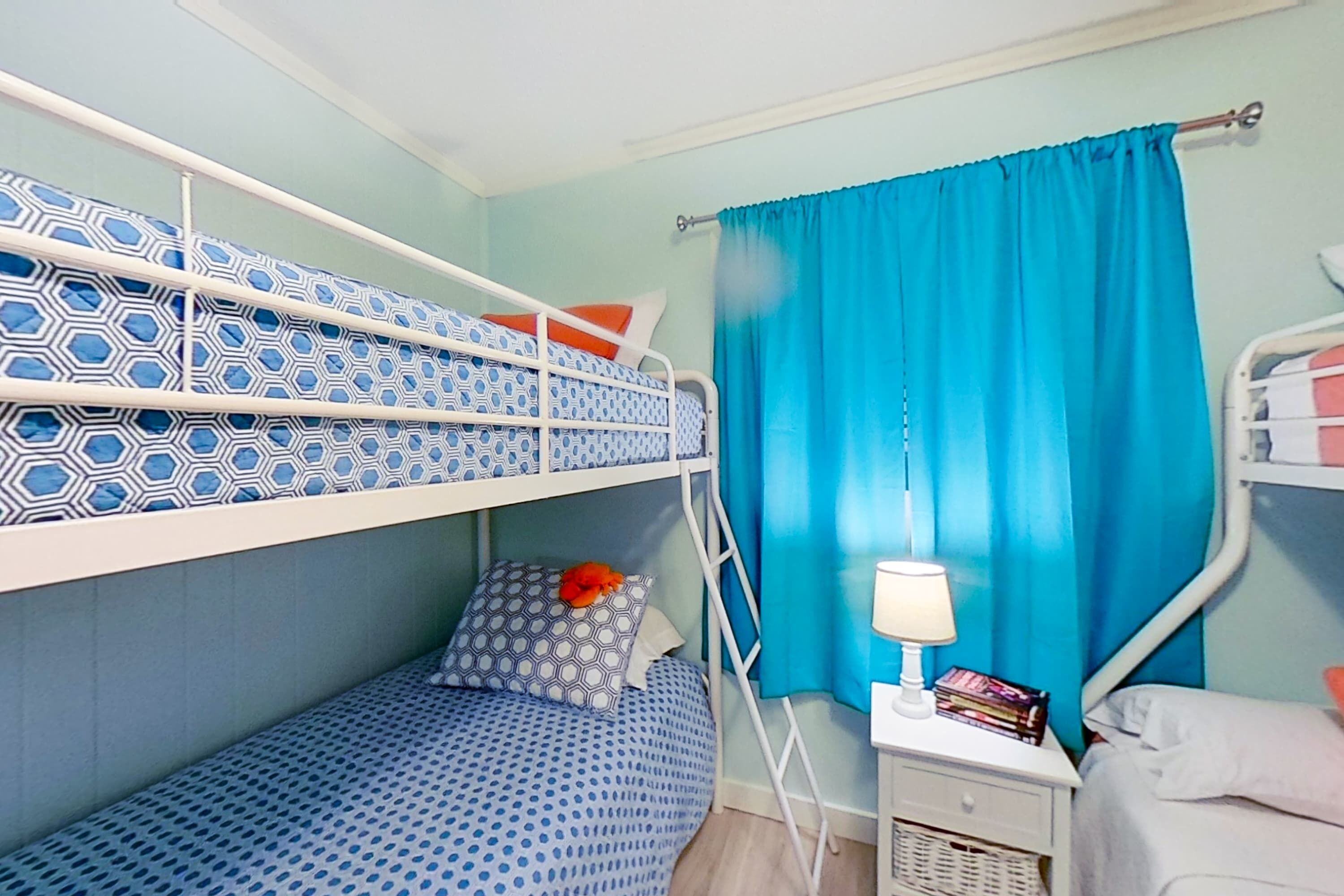 Une chambre avec un lit superposé, une couette à motifs, une table de chevet avec une lampe et une fenêtre avec des rideaux bleus.
