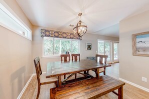A dining area with a wooden table and chairs, a chandelier, and large windows with a view of greenery. - Tree-lined home with beautiful patio, deck & firepit - near golf & the beach (Hilton Head)
