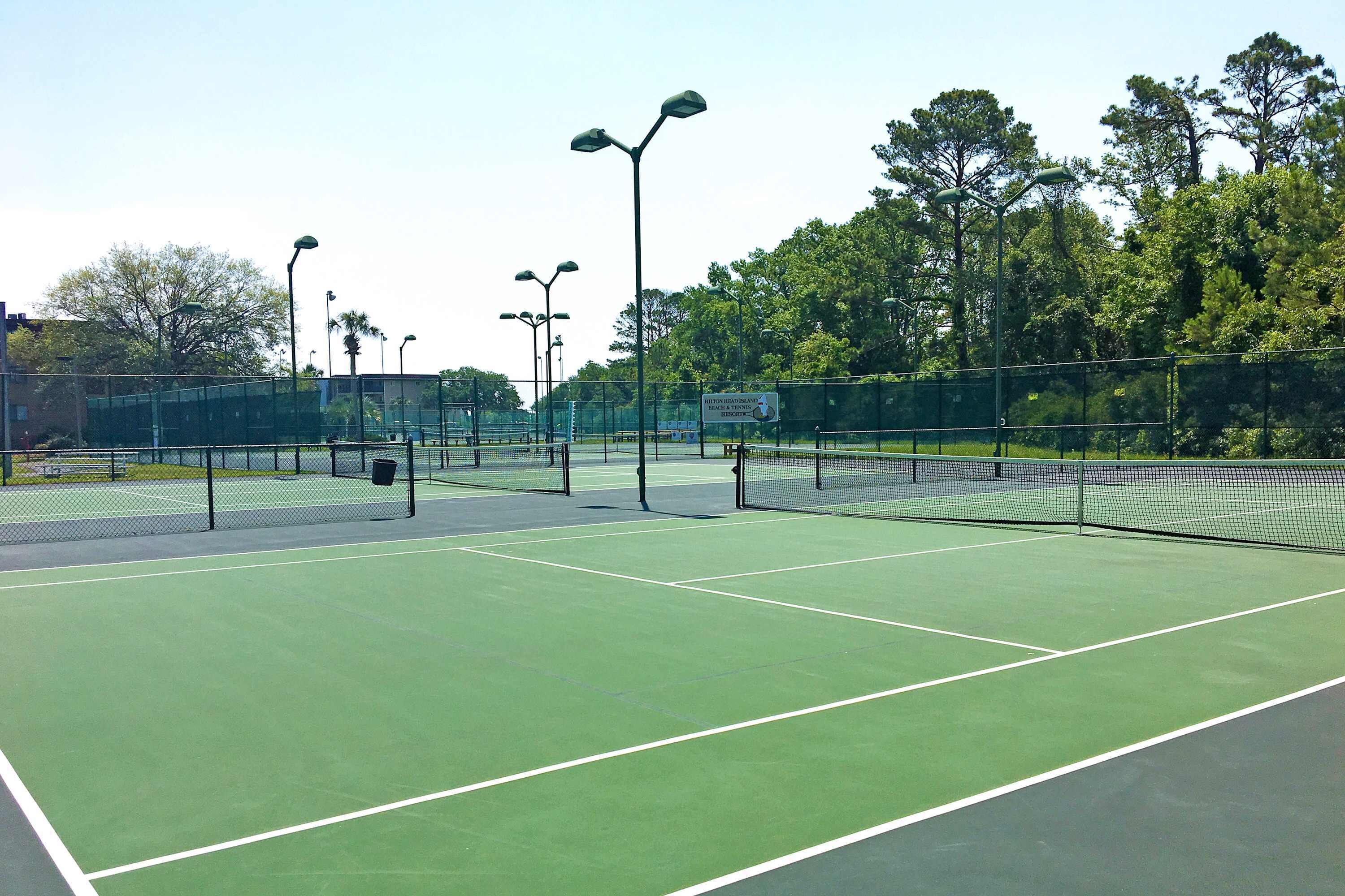 Un court de tennis comprenant plusieurs terrains, entouré d’arbres et de lampadaires.