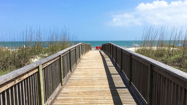 Ein hölzerner Steg führt zu einem Strand mit blauem Himmel und weiter Meerblick.