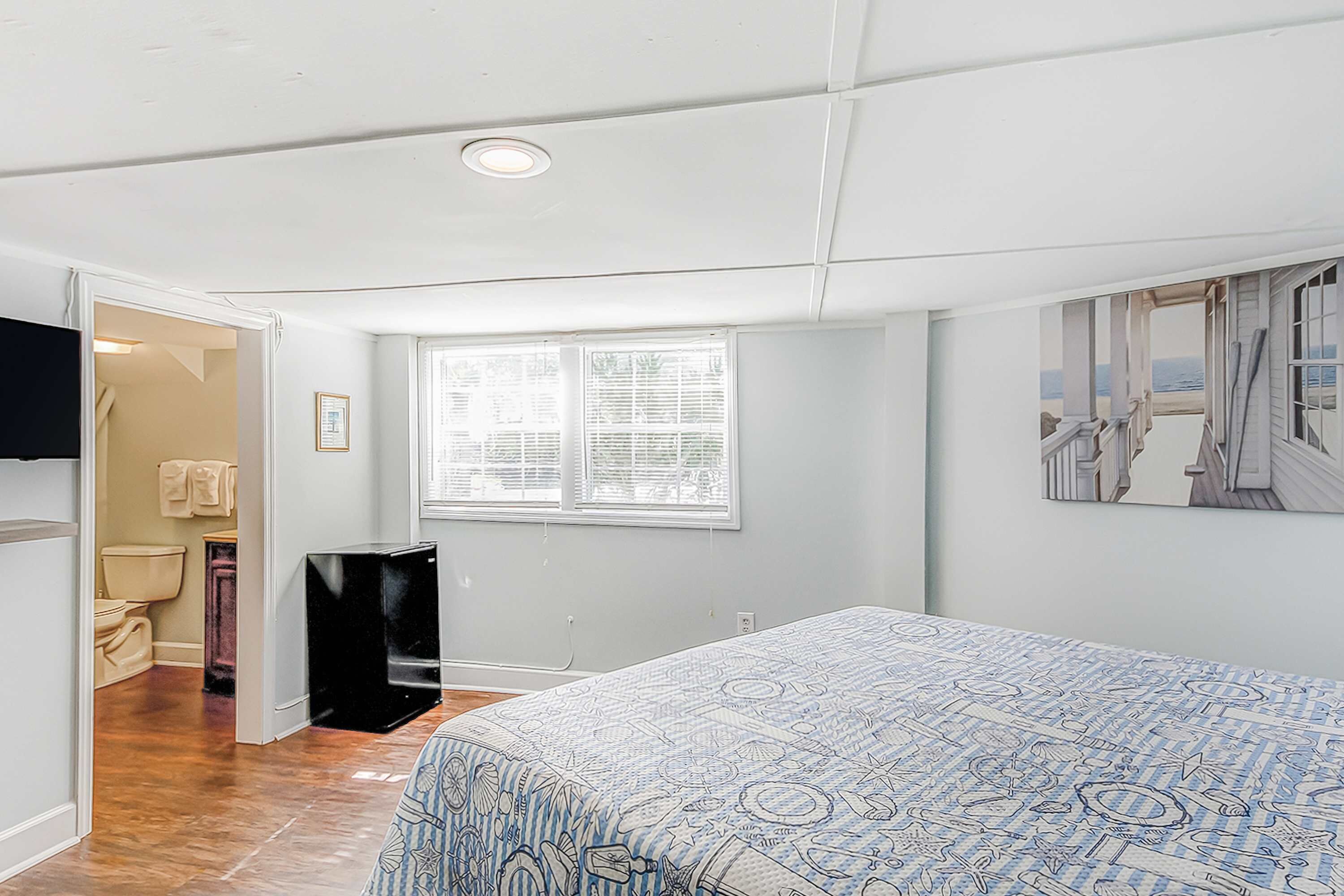 A bedroom with a bed, a window, a door leading to a bathroom, and a wall-mounted artwork.