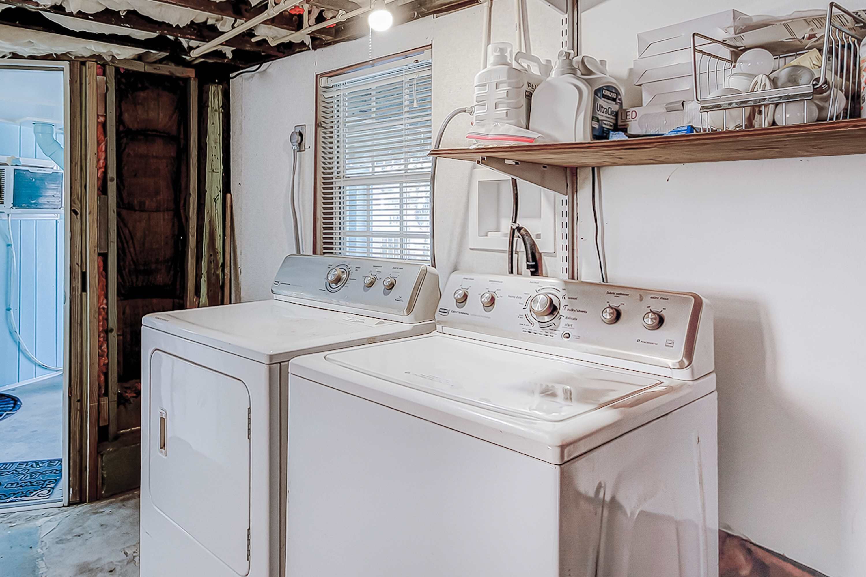 Une buanderie équipée de deux machines à laver et deux sèche-linge superposés, d’une étagère en bois où sont rangées des bouteilles, et d’une fenêtre avec des stores.
