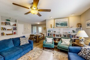 A living room with a blue sofa, teal armchairs, a ceiling fan, and a bookshelf. - Beach Around the Corner Quiet Home w/ 2 Screened Porches, WiFi & Laundry (Edisto Beach)