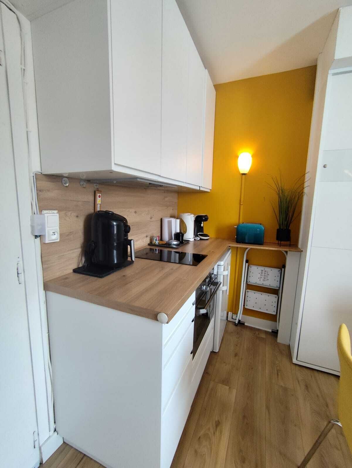 A compact kitchen with white cabinets, a wooden countertop, a coffee maker, a kettle, a toaster, and a small plant.