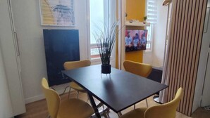A dining area with a table and chairs, a TV, and a window with blinds. - Le 4 Saisons (Ustou)