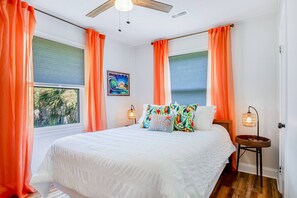 A bedroom with a bed, two windows with curtains, a ceiling fan, a wall-mounted lamp, and a painting. - Dreamy Coastal Oasis W/ Large Deck, Fast WiFi, Beach Access Nearby & Central AC (Edisto Beach)