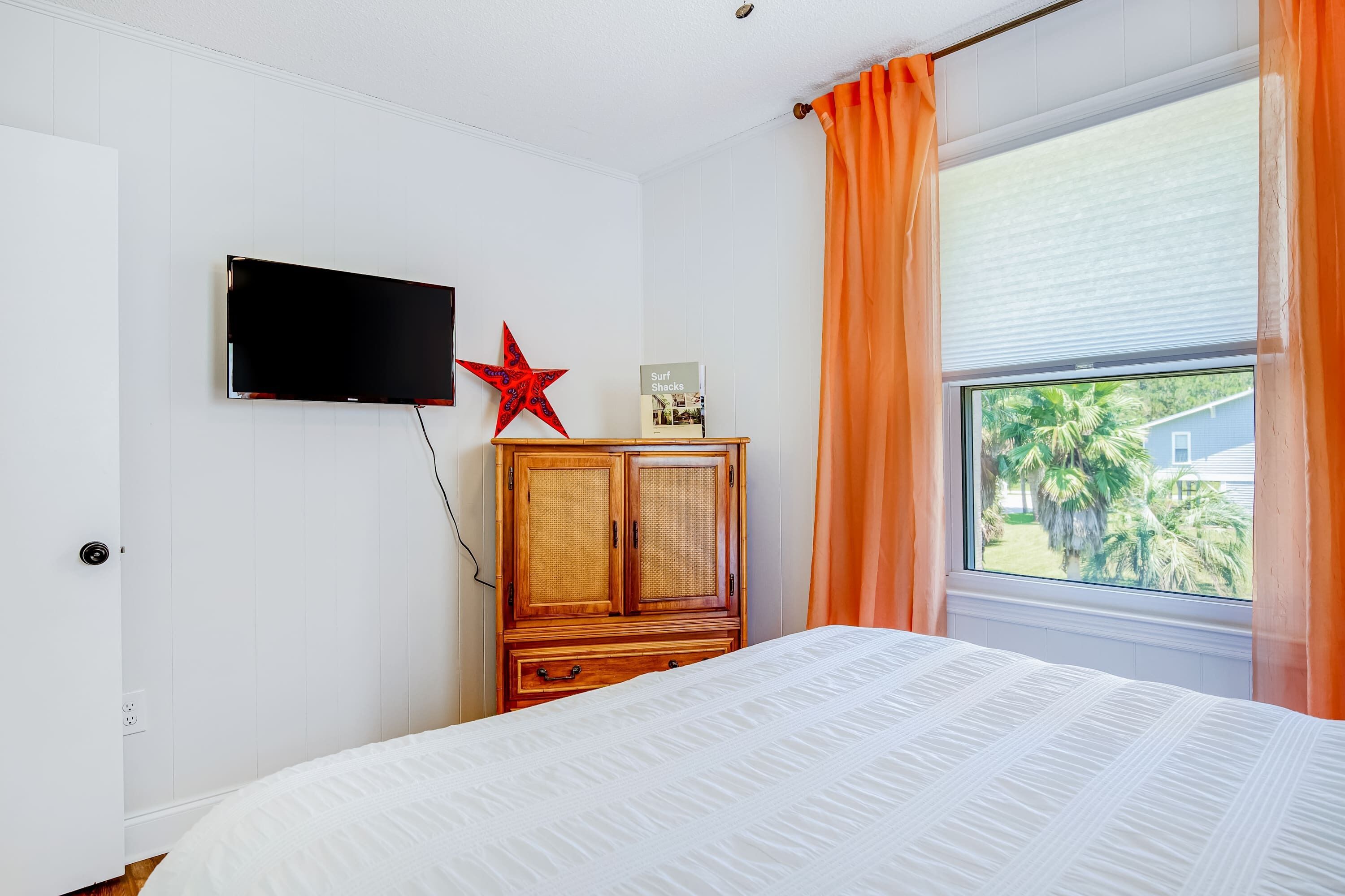 Un dormitorio con una cama, televisión montada en la pared, un armario de madera y una ventana con cortinas.