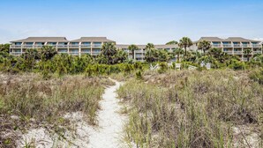 A beachfront hotel with multiple balconies, a sandy path leading to the building, and surrounding greenery. - Condo with Coligny Beach views, outdoor pool, beach access, balcony, WD, & AC (Hilton Head)