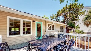 A covered patio with a metal table and chairs, a white picket fence, and a house with large windows and a teal door. - Ocean-view dog-friendly home across street from beach with private pool, W/D, AC (Edisto Beach)