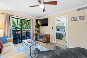 A living room with a sofa, coffee table, and a TV. There's a balcony with outdoor furniture and a ceiling fan. - Coastal condo with balcony, washer/dryer, heated pool, hot tub, & tennis courts (Hilton Head)