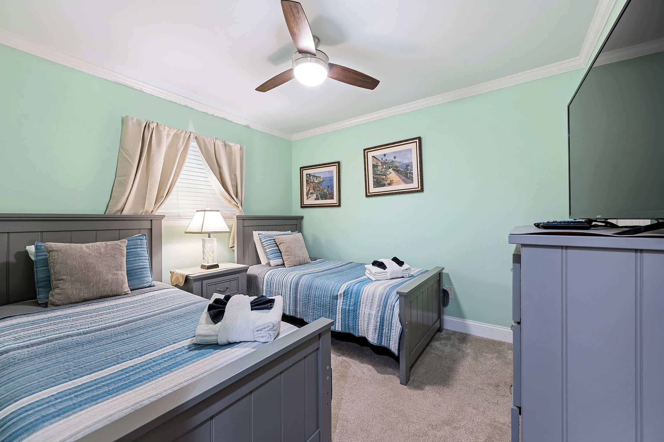 A bedroom with two beds, a ceiling fan, a desk, and two framed pictures on the wall.