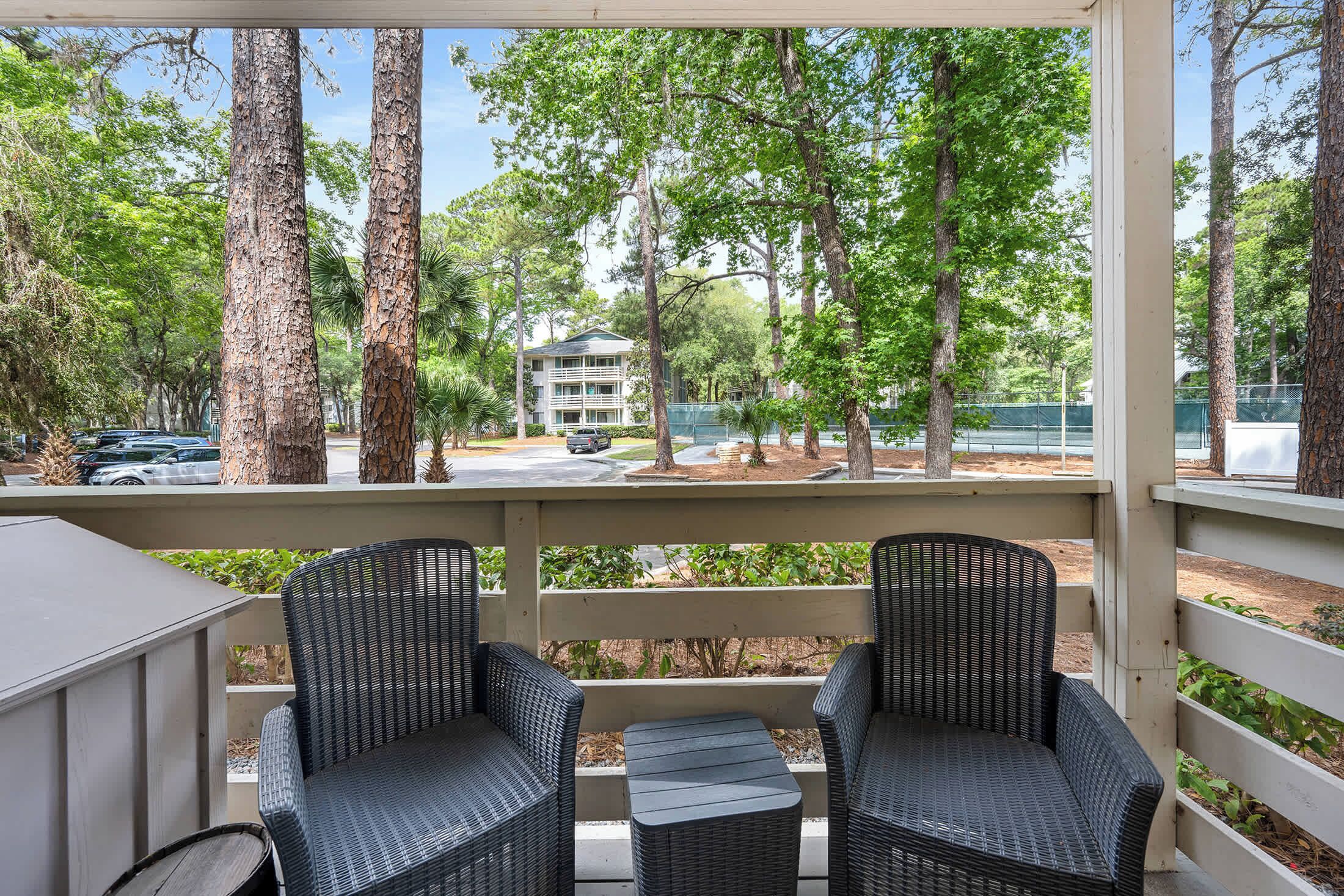 A covered outdoor seating area with wicker chairs and a table, overlooking a parking lot and trees.