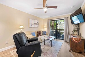 A living room with a sofa, a black leather chair, a coffee table, a television, and a balcony with a view of a building and trees. - Coastal condo with balcony, washer/dryer, heated pool, hot tub, & tennis courts (Hilton Head)