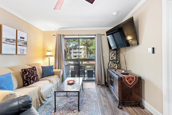 A living room with a sofa, coffee table, TV, and a view to the outside. - Coastal condo with balcony, washer/dryer, heated pool, hot tub, & tennis courts (Hilton Head)