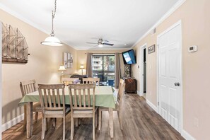 A dining area with wooden chairs and a table, a ceiling fan, a television, and a door. - Coastal condo with balcony, washer/dryer, heated pool, hot tub, & tennis courts (Hilton Head)
