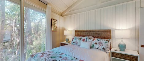 Un dormitorio con una cama grande, una mesita de noche con lámpara, un cabecero de mimbre y vistas a árboles a través de una puerta corrediza.