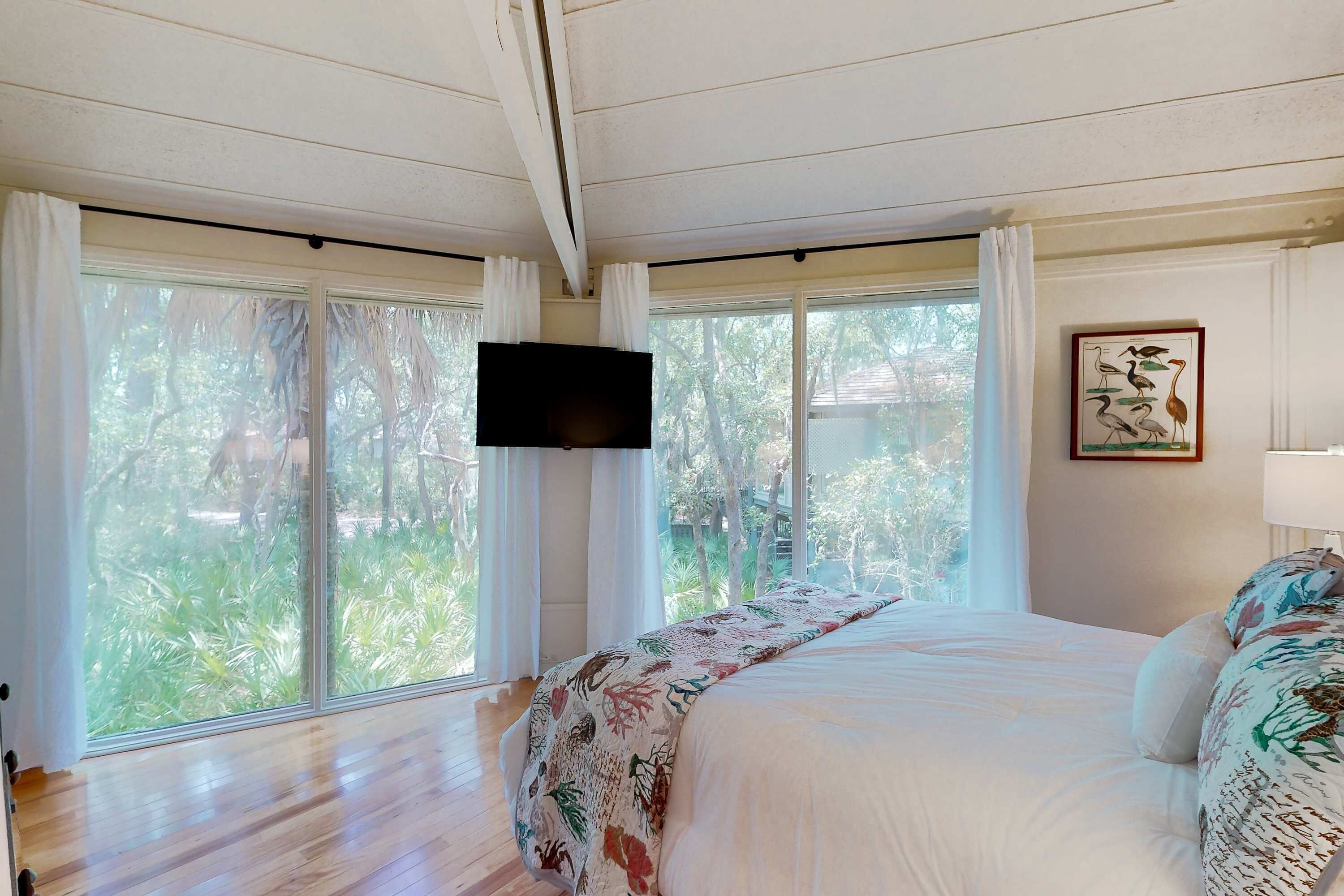 A bedroom with a bed, a flat-screen TV, large windows, and a framed picture on the wall.