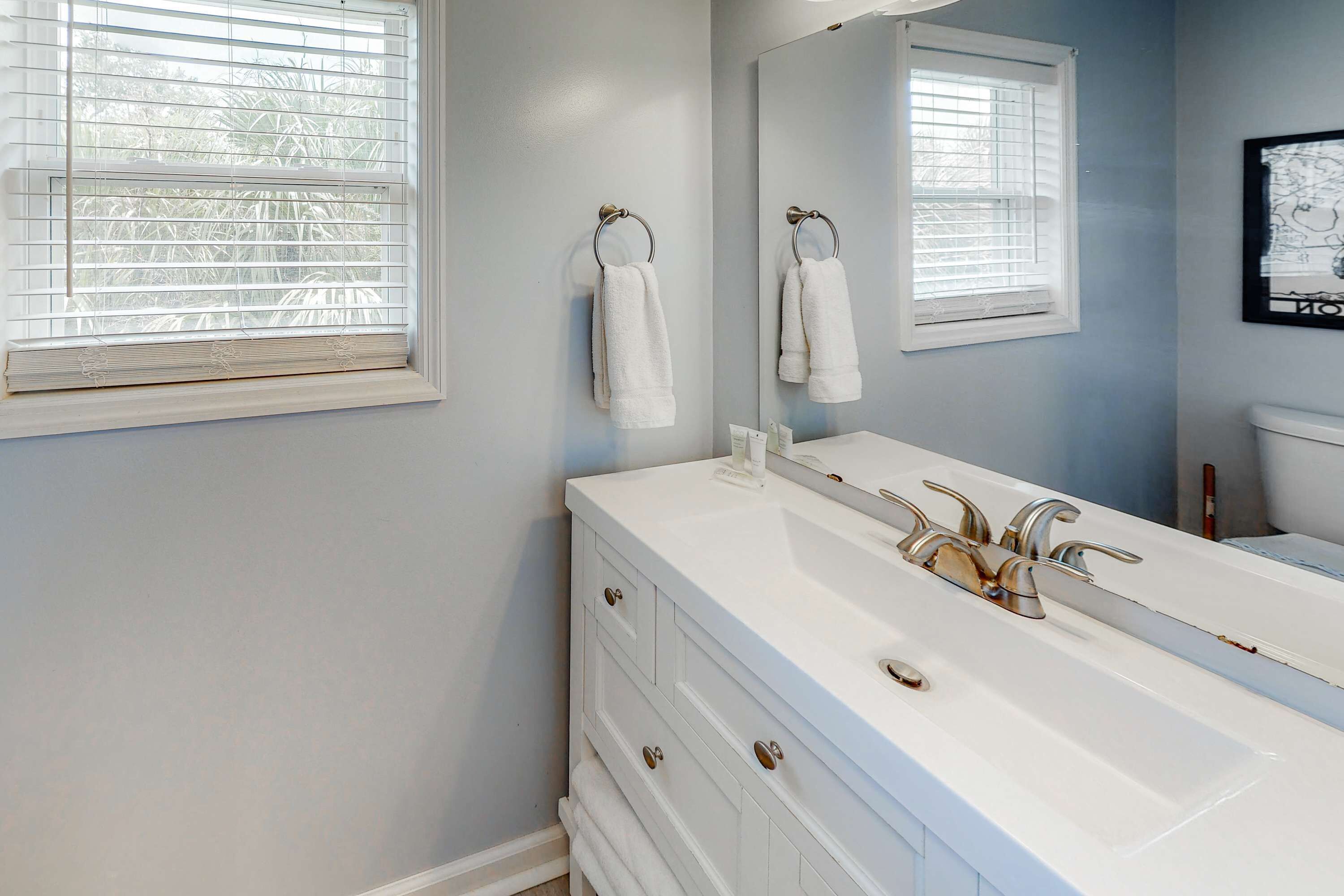A bathroom with a white vanity, a large mirror, and two windows with blinds.