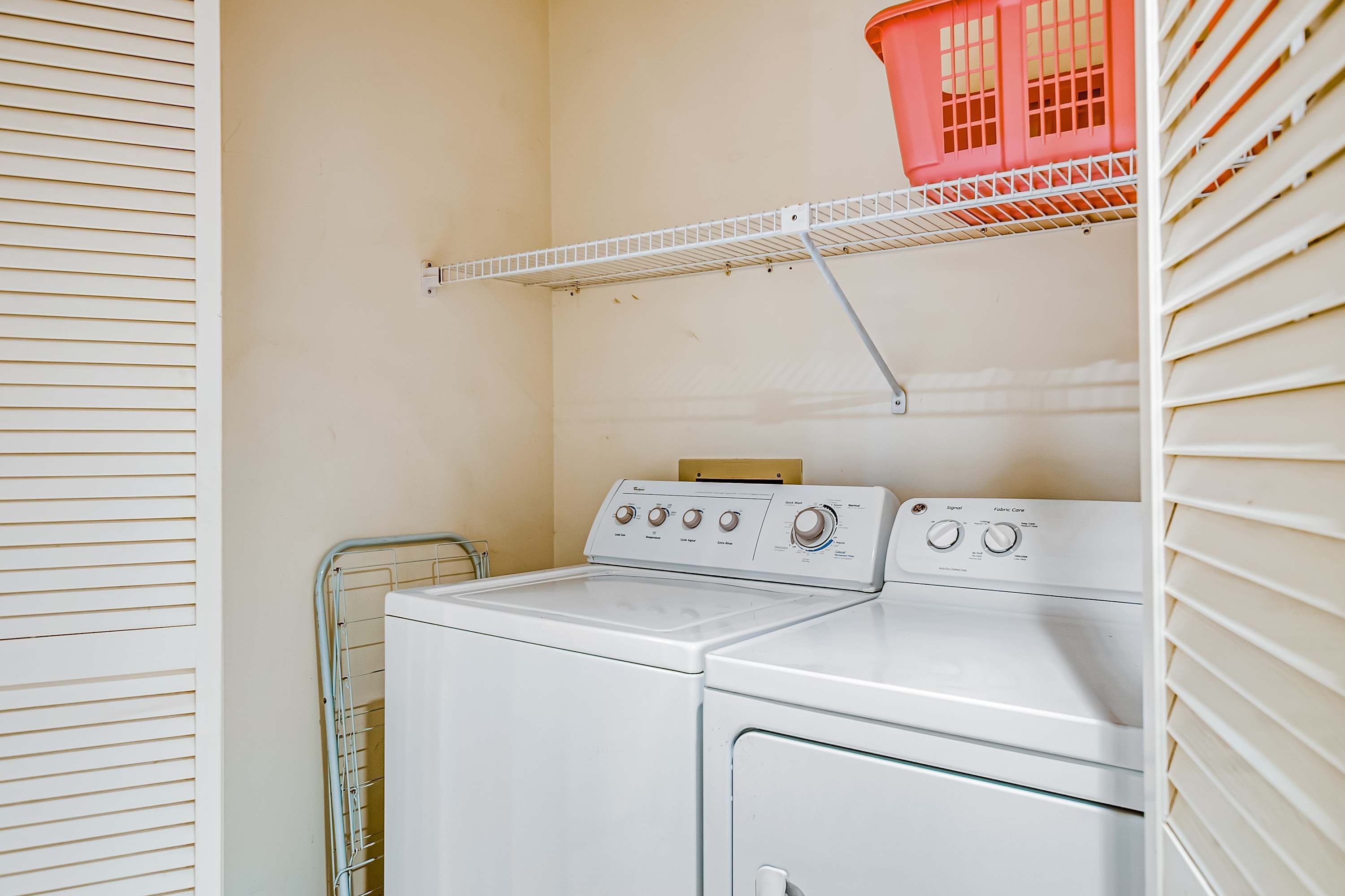 Un espace buanderie avec un lave-linge et un sèche-linge blancs, un étendoir métallique et un panier à linge rouge posé sur une étagère.