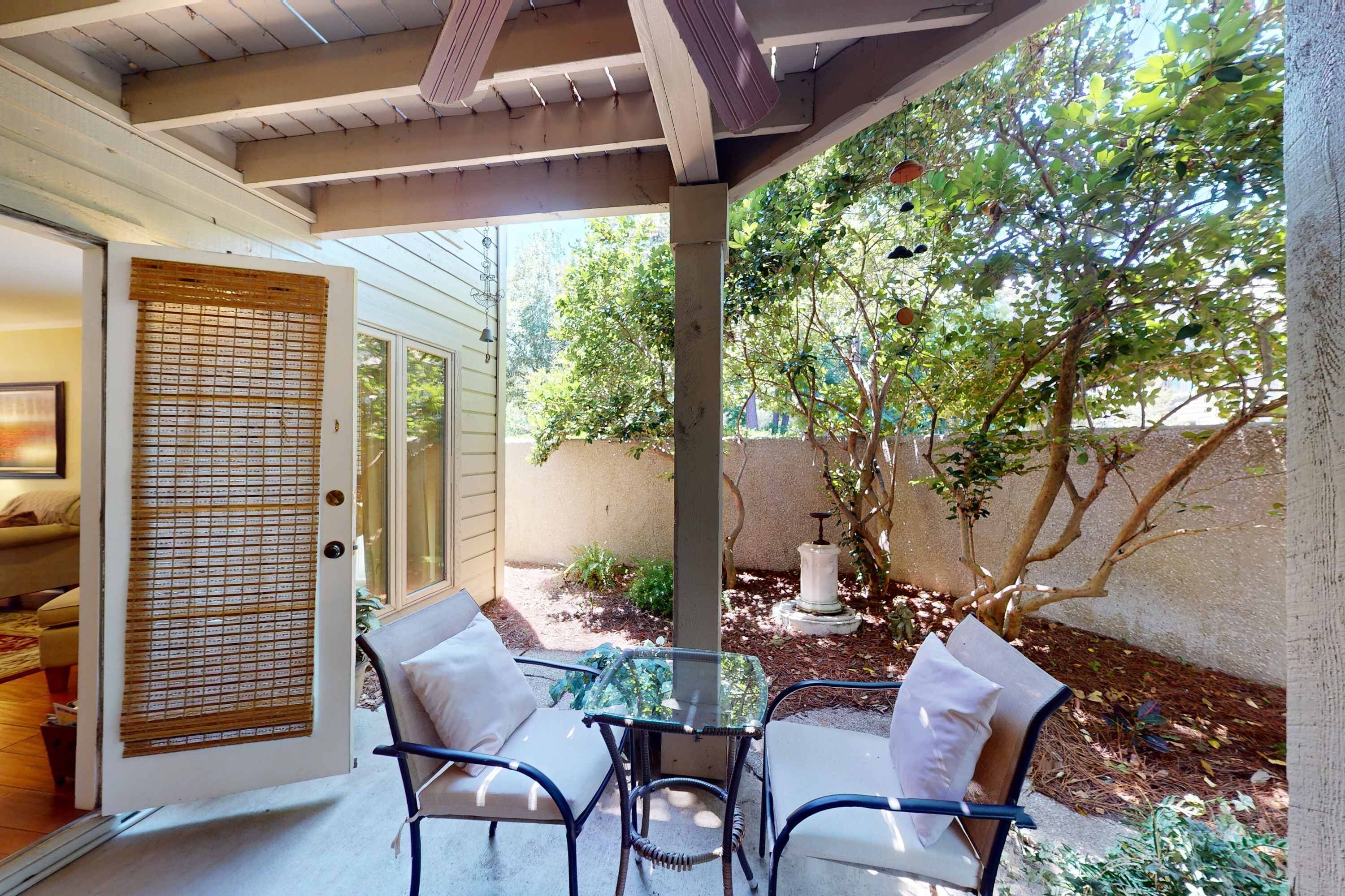 A covered patio with a glass table and chairs, a door with a bamboo screen, and a view of a garden with trees and a fountain.