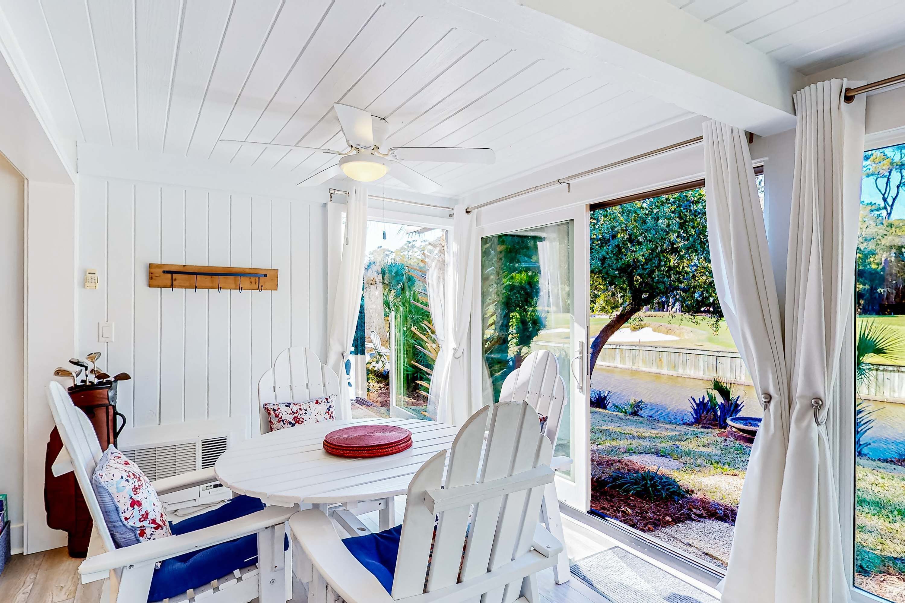 A sunlit room with a white ceiling fan, a wooden coat rack, a table with chairs, and a view of a garden through large windows.
