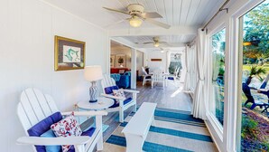 A room with white walls, a ceiling fan, a large window with a view of greenery, a blue and white striped rug, and a white bench.