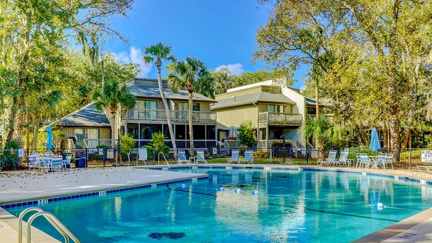 A swimming pool with clear blue water, surrounded by lounge chairs and umbrellas, with a building featuring balconies in the background. - Beautifully remodeled villa with WiFi, inviting porch & balconies walk to beach (Hilton Head)