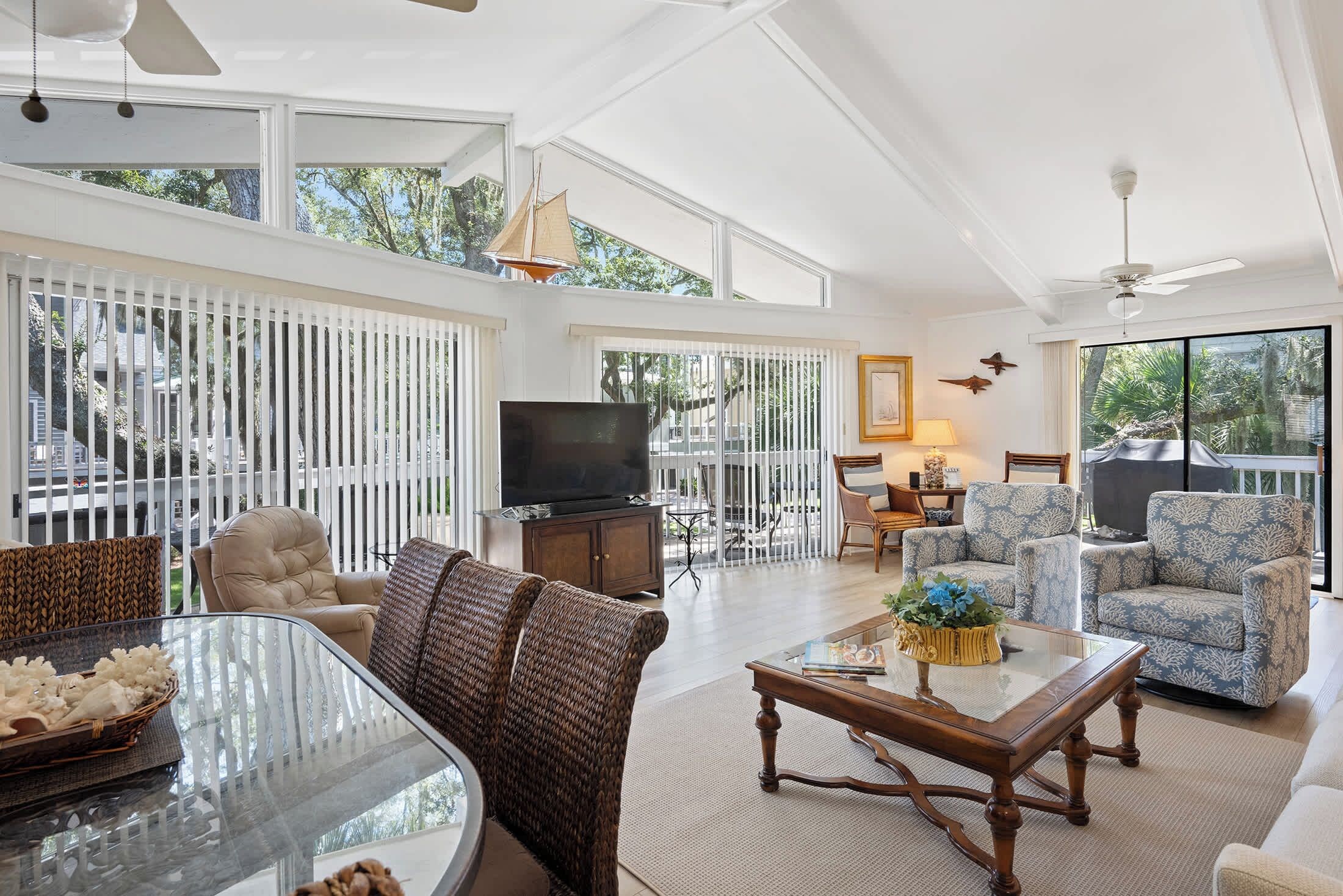 A spacious living room with a large glass table, comfortable seating, a television, and a view of the outdoors.