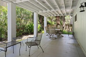 A covered patio area with metal chairs and tables, surrounded by lush greenery and a wooden fence. - North Forest Beach home with large deck, three houses from the beach (Hilton Head)