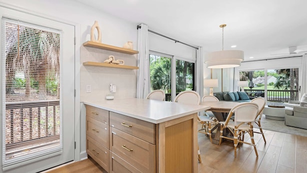 A modern kitchen with a central island, wooden cabinets, and a dining area with wicker chairs. - Scenic Golf Front Condo with Large Deck, Resort Pool & Easy Beach Access (Hilton Head)