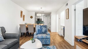 Een moderne woonkamer met een grijze bank, een fauteuil met een patroon, een eethoek met tafel en stoelen, en een keuken op de achtergrond.