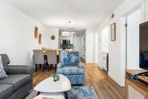 A modern living room with a grey sofa, a patterned armchair, a dining area with a table and chairs, and a kitchen in the background. - Family-friendly condo with top-floor balcony, complex pools, tennis, pickleball (Hilton Head)