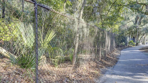 A paved pathway flanked by chain-link fences and dense foliage. - Bright and beautiful condo, 5 min to beach, golf and Van De Meer Tennis Courts (Hilton Head)