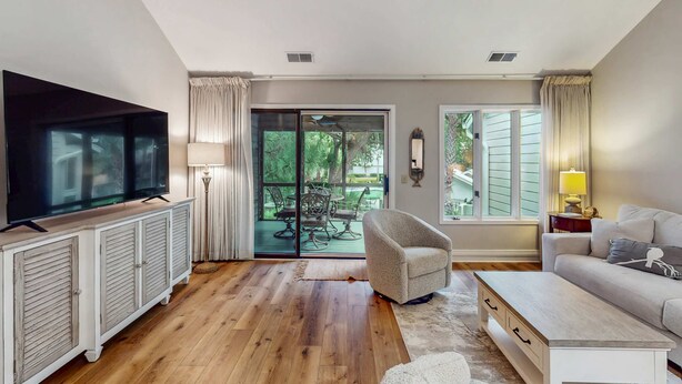 A living room with a flat-screen TV, a sofa, a coffee table, and a sliding glass door leading to an outdoor patio. - Pool view condo with screened in patio, complex pool hot tub, near golf (Hilton Head)