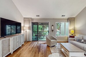 A living room with a flat-screen TV, a sofa, a coffee table, and a sliding glass door leading to an outdoor patio. - Pool view condo with screened in patio, complex pool hot tub, near golf (Hilton Head)