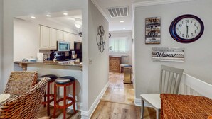 A kitchen with white cabinets, a dining area with a wicker chair and stools, and a wall clock. - Pool view condo with screened in patio, complex pool hot tub, near golf (Hilton Head)