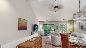 A modern living room with a television, wicker furniture, a dining table with placemats, and a large window with a view. - Pool view condo with screened in patio, complex pool hot tub, near golf (Hilton Head)