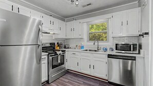 Une cuisine avec des meubles blancs, des appareils en acier inoxydable, une fenêtre avec des stores et un plancher en bois.