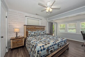 A bedroom with a bed, a nightstand, a lamp, a ceiling fan, and a window with blinds. - Escape to the serene beauty of the Smoky Mountains, Shiner's Hollow. (Gatlinburg)