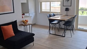 A modern living room with a black sofa, a dining table with chairs, and a large window providing natural light. - Divine Apartment by TOK (Durrës)
