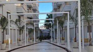 A covered walkway with potted plants and benches, leading to a building with balconies. - Divine Apartment by TOK (Durrës)